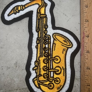 May include: A gold embroidered patch of a saxophone with a white outline and black backing.