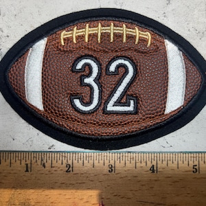 Pode incluir: Um remendo de futebol marrom e branco com o número 32 bordado em branco sobre um fundo preto.