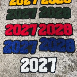 May include: A collection of chenille patches in various colors, including yellow, green, red, blue, and white. Each patch displays the numbers "2027" and "2028" in a bold, collegiate-style font, perfect for apparel customization.