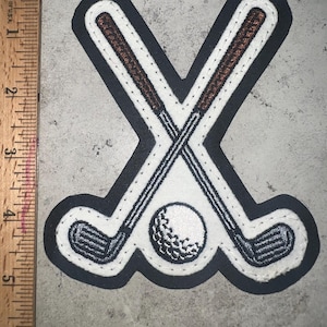 May include: Embroidered patch featuring two crossed golf clubs and a golf ball. The patch has a black border, white background, and brown club handles. The golf ball is white with detailed stitching.