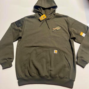 Puede incluir: Sudadera con capucha verde oliva con bolsillo de canguro y capucha con cord&oacute;n. Cuenta con un parche con el logotipo de un b&uacute;falo y un parche de la bandera estadounidense en la manga izquierda. Incluye una etiqueta Carhartt.