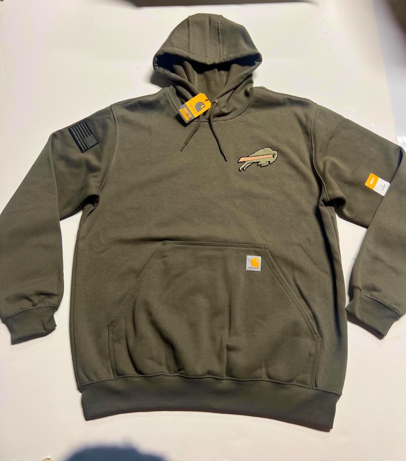 Puede incluir: Sudadera con capucha verde oliva con bolsillo de canguro y capucha con cord&oacute;n. Cuenta con un parche con el logotipo de un b&uacute;falo y un parche de la bandera estadounidense en la manga izquierda. Incluye una etiqueta Carhartt.