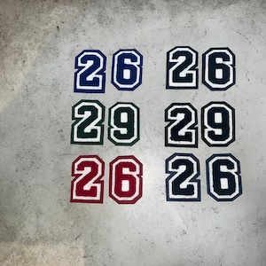 May include: A collection of fabric patches featuring the numbers 26 and 29 in various colors. The patches have a white outline and are in navy blue, green, red, and black. The numbers are in a block font.