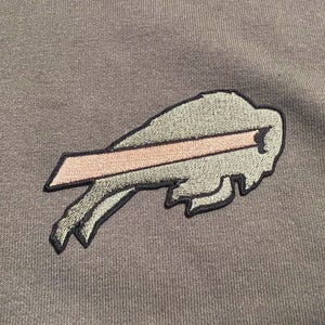 Puede incluir: Primer plano de una sudadera con capucha gris con un logotipo bordado de los Buffalo Bills. El logotipo es una cabeza de bisonte estilizada con una franja rosa, delineada en negro. El cord&oacute;n de la sudadera con capucha es visible.