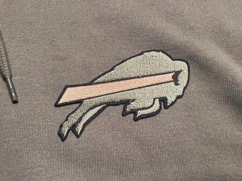 Puede incluir: Primer plano de una sudadera con capucha gris con un logotipo bordado de los Buffalo Bills. El logotipo es una cabeza de bisonte estilizada con una franja rosa, delineada en negro. El cord&oacute;n de la sudadera con capucha es visible.