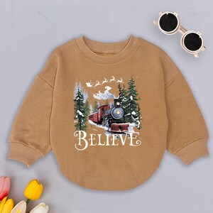 May include: A brown sweatshirt with a Christmas-themed design featuring a red train with white snow on top, surrounded by pine trees and the words "BELIEVE".