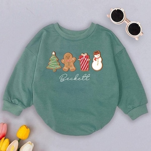 May include: A green long-sleeved sweatshirt with a Christmas theme. The sweatshirt features a gingerbread man, a Christmas tree, a present, and a snowman. The text "Beckett" is printed on the front of the sweatshirt.