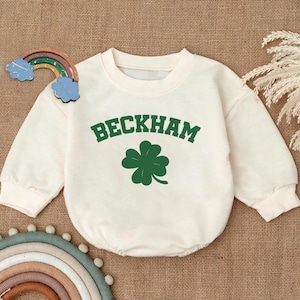 May include: A cream-colored long-sleeved sweatshirt with a green four-leaf clover and the text "BECKHAM" in green.