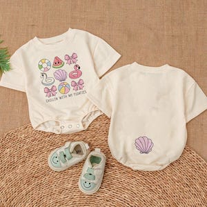 May include: Two white short-sleeved baby onesies. The onesie on the left has a watermelon, flamingo, and other summer-themed illustrations with the text "Chillin' with my floaties". The onesie on the right has a single seashell illustration.