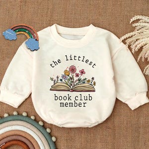 May include: A white long-sleeved onesie with the text "the littlest book club member" and a drawing of a book with flowers on top.