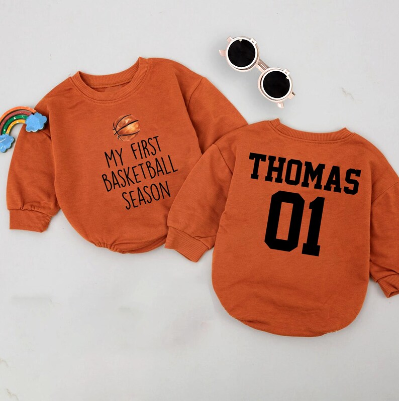 May include: Two orange long-sleeved shirts with a basketball theme. One shirt says "My First Basketball Season" with a basketball graphic. The other shirt says "Thomas 01".