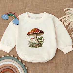May include: A white long-sleeved baby onesie with a mushroom graphic. The mushroom is red with white spots and has a green stem. There are smaller mushrooms around the base of the large mushroom.