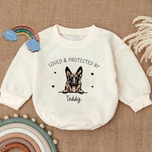 May include: Cream-colored baby onesie with long sleeves. The front features a German Shepherd graphic with the text "LOVED & PROTECTED BY" above and "Teddy" below. Small black hearts are scattered around the design.