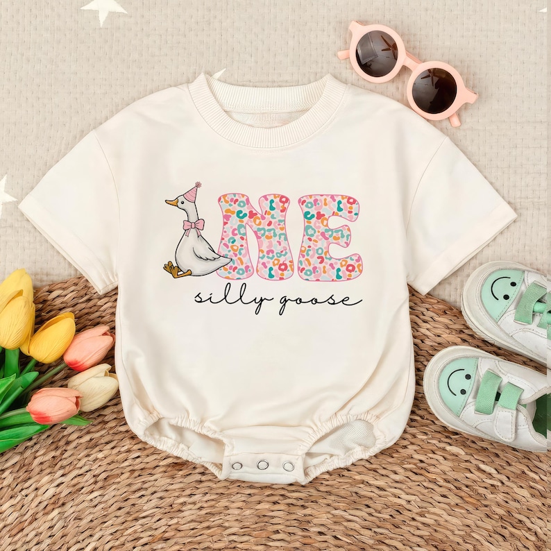 以下が含まれることがあります： ピンク、グリーン、ブルーの「ONE」が書かれた白いロンパース。帽子をかぶったガチョウが描かれています。「silly goose」という文字が「ONE」の下にあります。