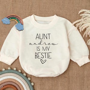 May include: A white long-sleeved onesie with the text "AUNT andrew IS MY BESTIE" and a small heart.