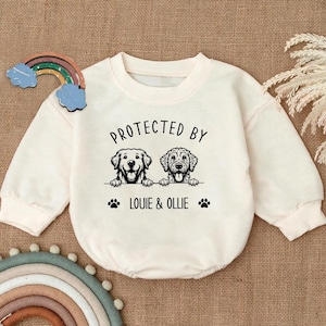 May include: A cream-colored long-sleeved baby onesie with a black graphic of two dogs peeking over a wall. The text "PROTECTED BY" is above the dogs and "LOUIE & OLLIE" is below the dogs.
