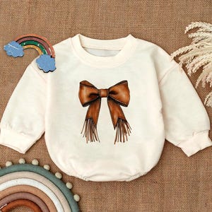May include: A white baby onesie with a brown bow with fringe detail. The bow is centered on the onesie.