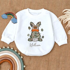 May include: White long-sleeved baby onesie with a camouflage bunny wearing a hat that says "Egg Hunter" and the name "William" printed below.
