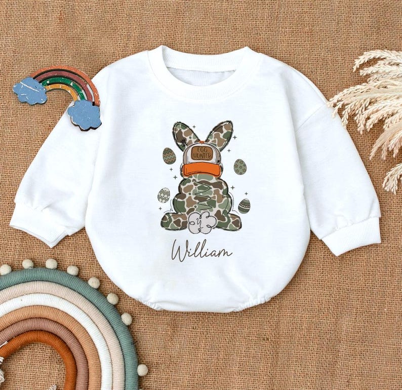 May include: White long-sleeved baby onesie with a camouflage bunny wearing a hat that says "Egg Hunter" and the name "William" printed below.