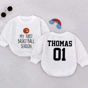 May include: Two white long-sleeved shirts. One shirt has a basketball graphic and the text "My First Basketball Season". The other shirt has the text "Thomas 01" in black.