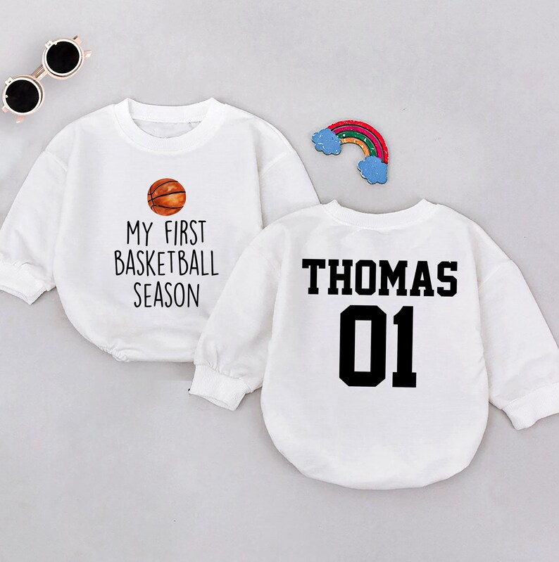 May include: Two white long-sleeved shirts. One shirt has a basketball graphic and the text "My First Basketball Season". The other shirt has the text "Thomas 01" in black.