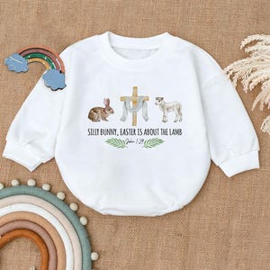 May include: White long-sleeve onesie with a watercolor illustration of a bunny, a cross, a lamb, and green leaves. The text reads "Silly Bunny, Easter is about the Lamb. John 1:29".