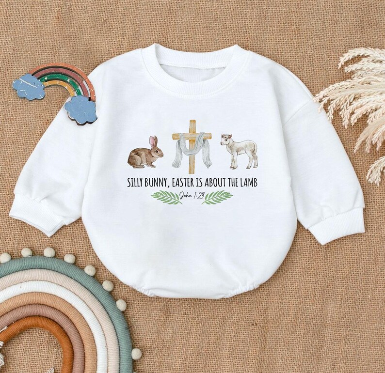 May include: White long-sleeve onesie with a watercolor illustration of a bunny, a cross, a lamb, and green leaves. The text reads "Silly Bunny, Easter is about the Lamb. John 1:29".
