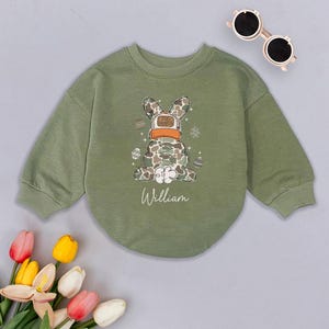 May include: Green long-sleeved sweatshirt with a camouflage bunny rabbit wearing a hat that says "Egg Hunter" and surrounded by Easter eggs. The sweatshirt has the name "William" printed on the front.