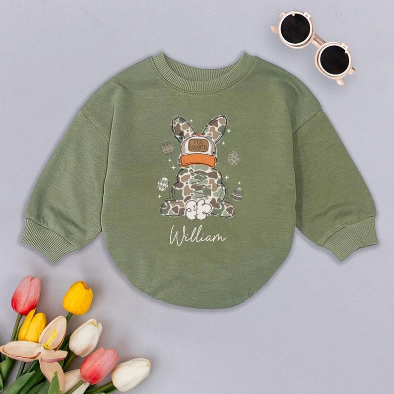 May include: Green long-sleeved sweatshirt with a camouflage bunny rabbit wearing a hat that says "Egg Hunter" and surrounded by Easter eggs. The sweatshirt has the name "William" printed on the front.