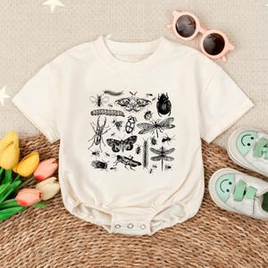May include: A cream-colored short-sleeve baby bodysuit featuring a black ink-style graphic print of various insects, including beetles, moths, and dragonflies. The garment has a round neckline and snap closures at the bottom for easy access.