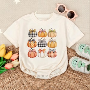 May include: Cream-colored baby bodysuit featuring a grid of nine illustrated pumpkins. Each pumpkin has a different pattern and bow, including gingham, polka dots, and stripes. The bows are in various colors, including pink, orange, and green.