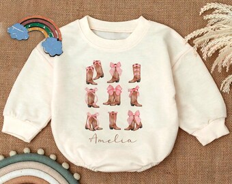 May include: A cream-colored sweatshirt with a graphic of pink bows and brown cowboy boots. The text "Amelia" is printed below the graphic.