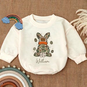May include: A white long-sleeved baby onesie with a camouflage Easter bunny wearing a hat that says "Egg Hunter" and the name "William" printed below the bunny.