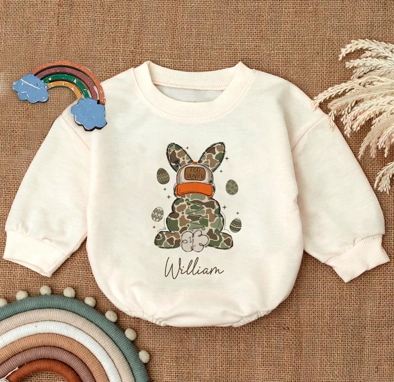 May include: A white long-sleeved baby onesie with a camouflage Easter bunny wearing a hat that says "Egg Hunter" and the name "William" printed below the bunny.