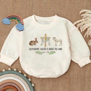 May include: A white long-sleeved onesie with a graphic of a bunny, a cross, a lamb, and the text "Silly Bunny, Easter is about the Lamb" with the scripture reference "John 1:29".