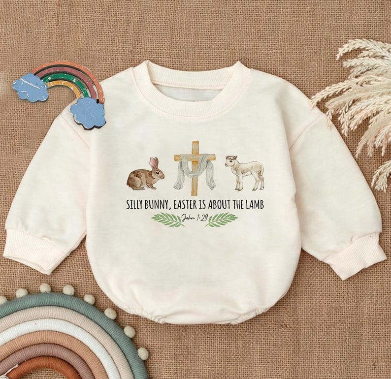 May include: A white long-sleeved onesie with a graphic of a bunny, a cross, a lamb, and the text "Silly Bunny, Easter is about the Lamb" with the scripture reference "John 1:29".