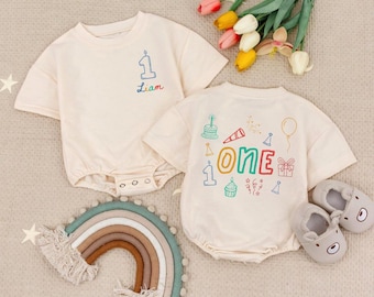 Op de afbeelding: Twee crèmekleurige babyrompers met kleurrijke geborduurde ontwerpen. Eén zegt "Liam" en "1", de andere zegt "ONE" met verjaardagsafbeeldingen. Een regenboog wandkleed en een paar grijze babyschoentjes in berenvorm staan ook in de afbeelding.