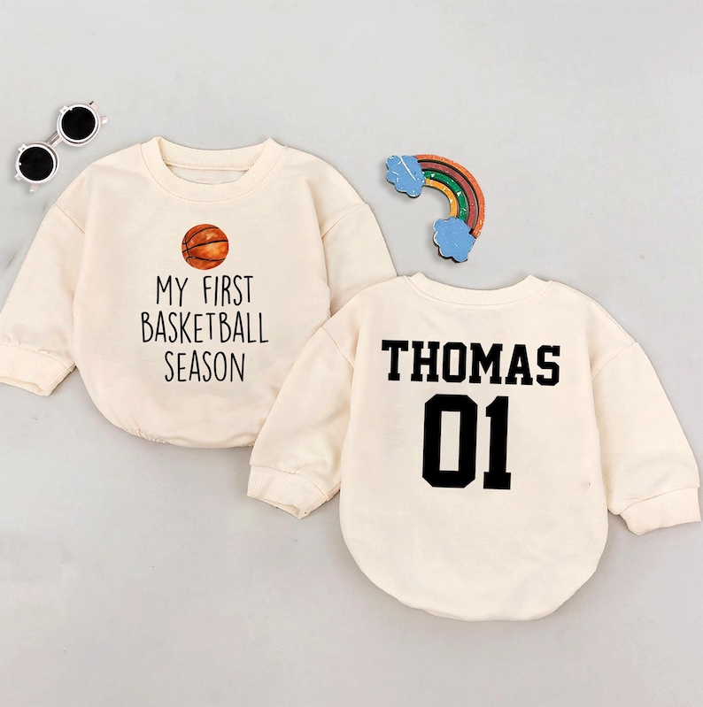 May include: Two cream-colored long-sleeved shirts with a basketball theme. One shirt says "My First Basketball Season" with a basketball graphic. The other shirt says "Thomas 01" on the back.