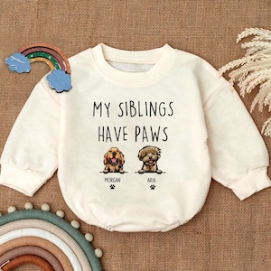 May include: A white baby onesie with the text "My siblings have paws" and two illustrations of dogs. The dogs are labeled "Morgan" and "Aria".