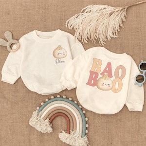 May include: Two cream-colored baby rompers with long sleeves. One features a dumpling graphic and the name "Olivia". The other has "BAO BOO" text and a dumpling graphic. A rainbow, sunglasses, and a bunny teether are also in the image.