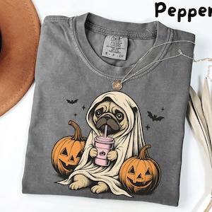 Puede incluir: Una camiseta gris Comfort Colors con un diseño de Halloween. El diseño presenta un pug vestido de fantasma, sosteniendo una bebida rosa, con dos calabazas talladas y murciélagos. La palabra "Pepper" está impresa en la parte superior.