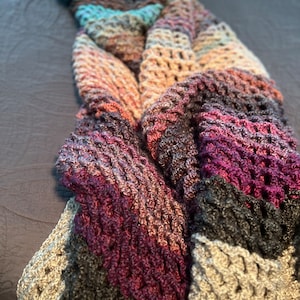 May include: A multi-colored crocheted blanket with a textured pattern. The blanket is made with shades of purple, pink, gray, and blue.