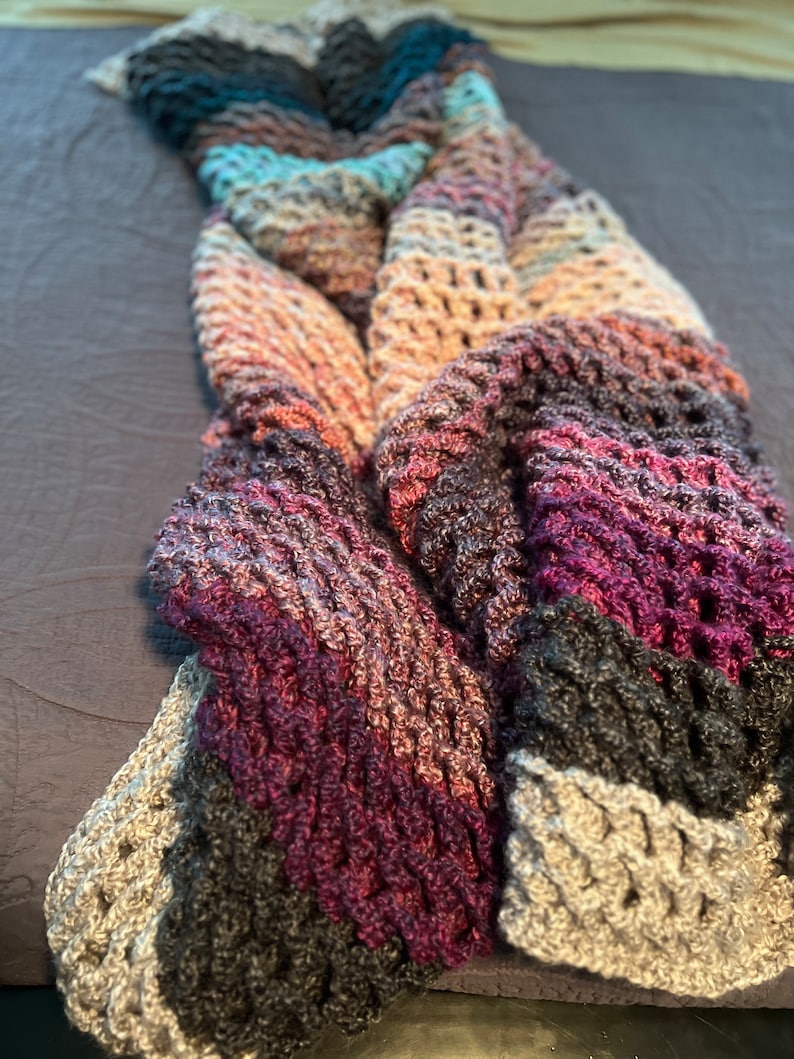 May include: A multi-colored crocheted blanket with a textured pattern. The blanket is made with shades of purple, pink, gray, and blue.
