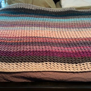 May include: A multi-colored crocheted throw blanket with a textured pattern. The blanket is made with a variety of shades of brown, pink, purple, and blue yarn.