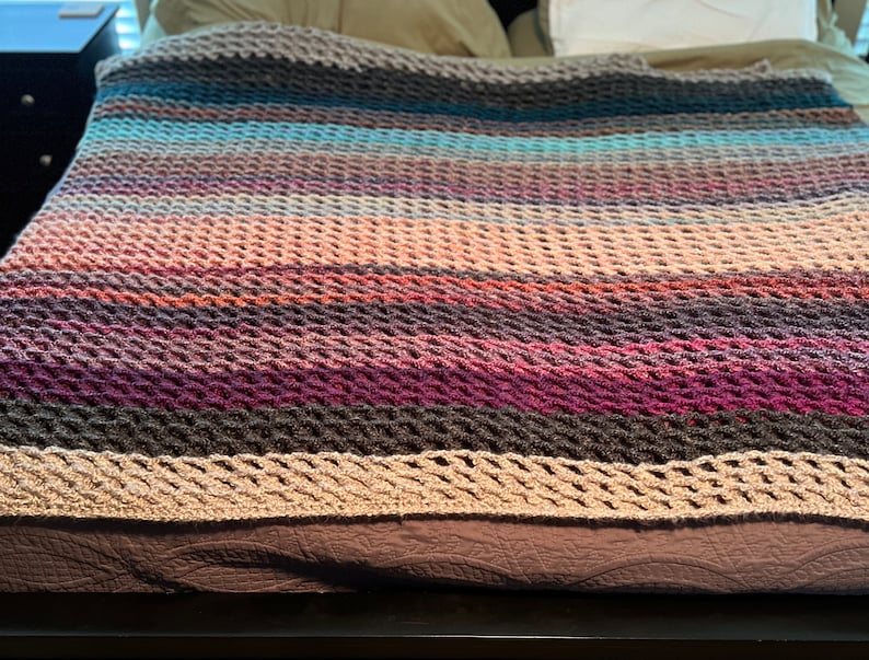 May include: A multi-colored crocheted throw blanket with a textured pattern. The blanket is made with a variety of shades of brown, pink, purple, and blue yarn.
