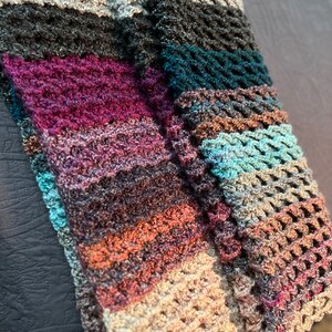 May include: A multi-colored crocheted scarf with a variety of textures and patterns. The scarf is made with a mix of light and dark colors, including beige, brown, teal, and purple.
