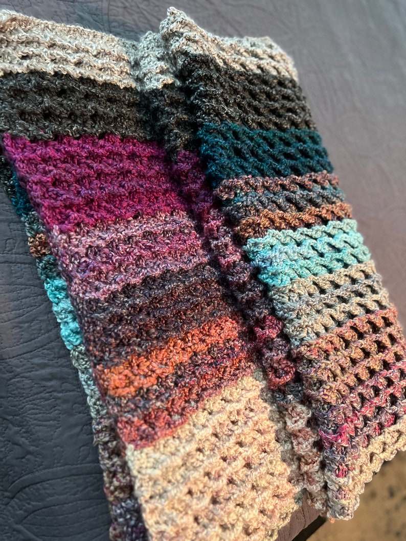 May include: A multi-colored crocheted scarf with a variety of textures and patterns. The scarf is made with a mix of light and dark colors, including beige, brown, teal, and purple.