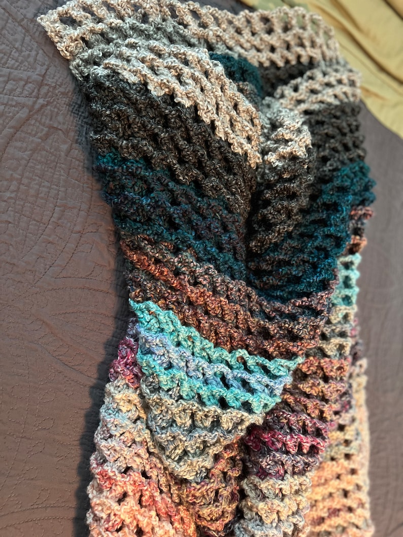 May include: A multi-colored crocheted blanket with a textured pattern. The blanket is made with a variety of shades of blue, brown, gray, and pink yarn.