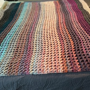 May include: A multi-colored crocheted blanket with a textured, honeycomb pattern. The blanket is made with a variety of shades of brown, blue, and purple yarn.