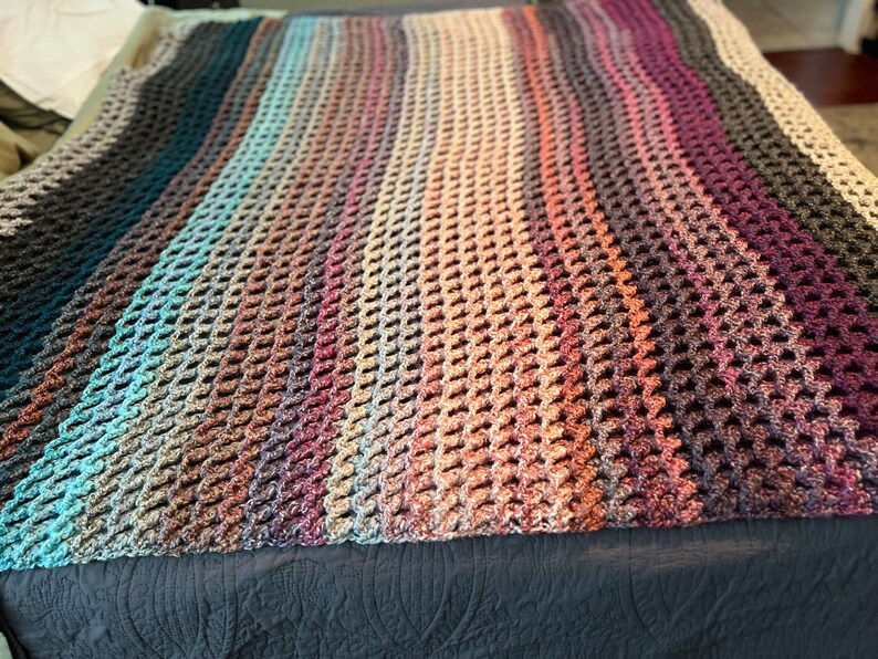May include: A multi-colored crocheted blanket with a textured, honeycomb pattern. The blanket is made with a variety of shades of brown, blue, and purple yarn.